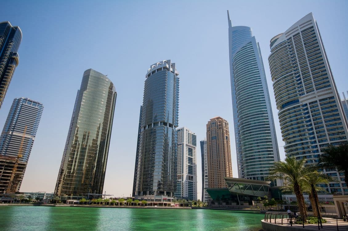 Skyline of JLT with glass facades reflecting sunlight, towering over a greenish body of water, under a clear blue sky.