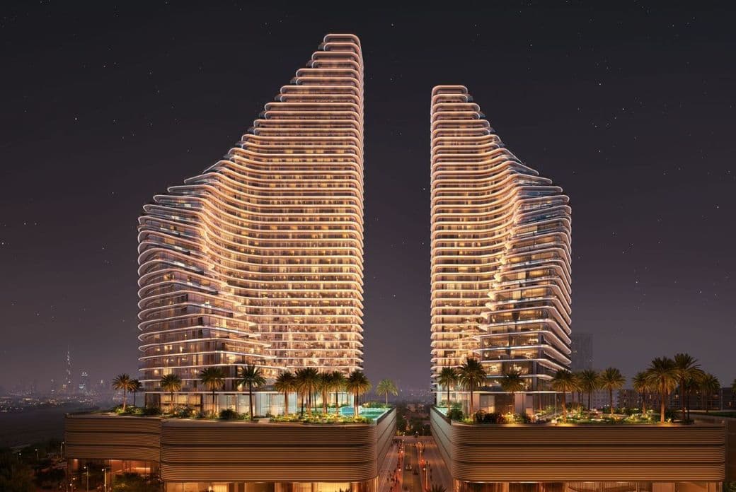 Nighttime view of Binghatti Flare showing two illuminated residential towers with curved architectural lines, landscaped podium areas, palm trees, and a visible city skyline in the background.
