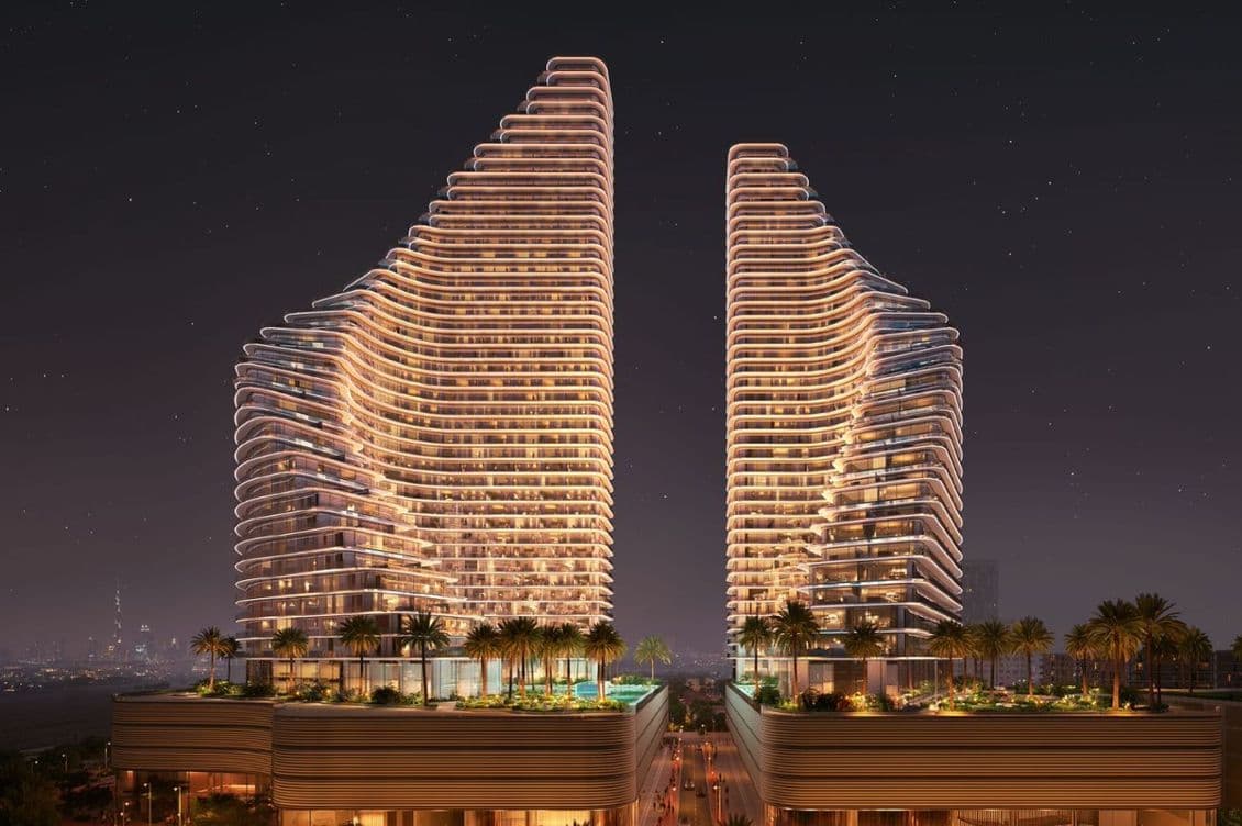 Nighttime view of Binghatti Flare showing two illuminated residential towers with curved architectural lines, landscaped podium areas, palm trees, and a visible city skyline in the background.