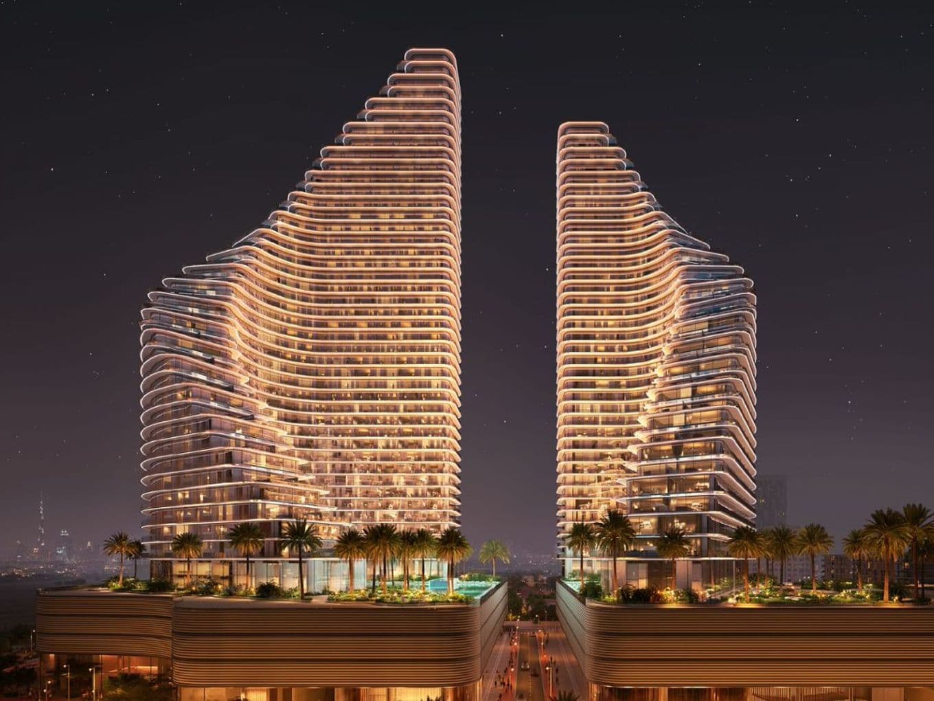 Nighttime view of Binghatti Flare showing two illuminated residential towers with curved architectural lines, landscaped podium areas, palm trees, and a visible city skyline in the background.
