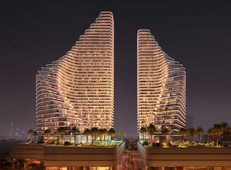 Nighttime view of Binghatti Flare showing two illuminated residential towers with curved architectural lines, landscaped podium areas, palm trees, and a visible city skyline in the background.