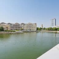 Waterfront view of beige buildings and a modern skyscraper under a clear blue sky. A lake reflects the buildings.