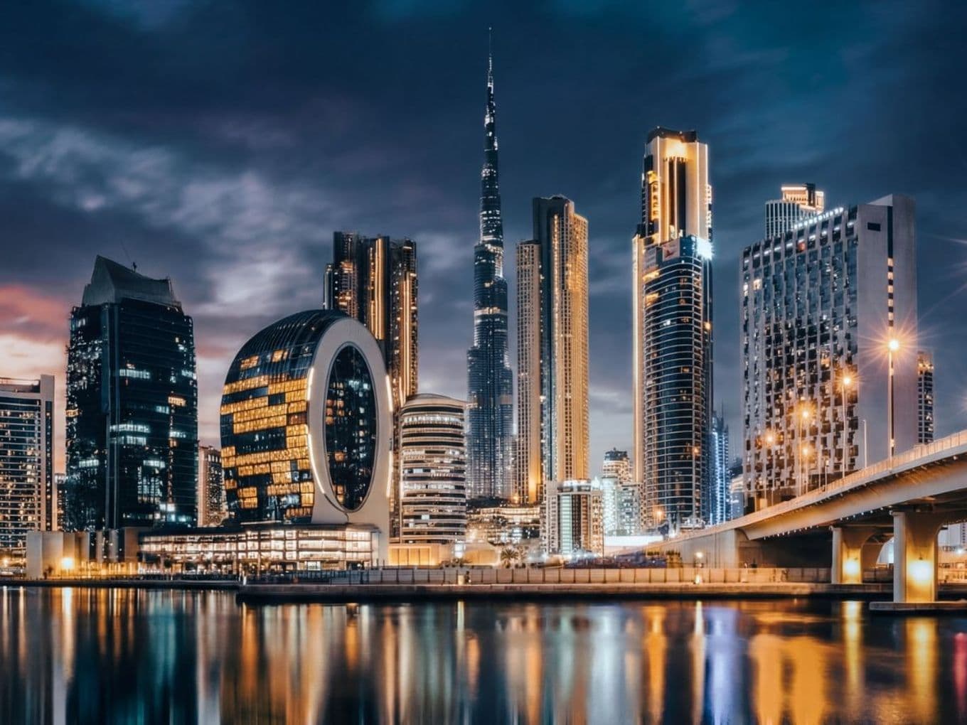 Dubai skyline at night with illuminated skyscrapers, including a distinctive circular building, reflected in calm water under a cloudy sky.