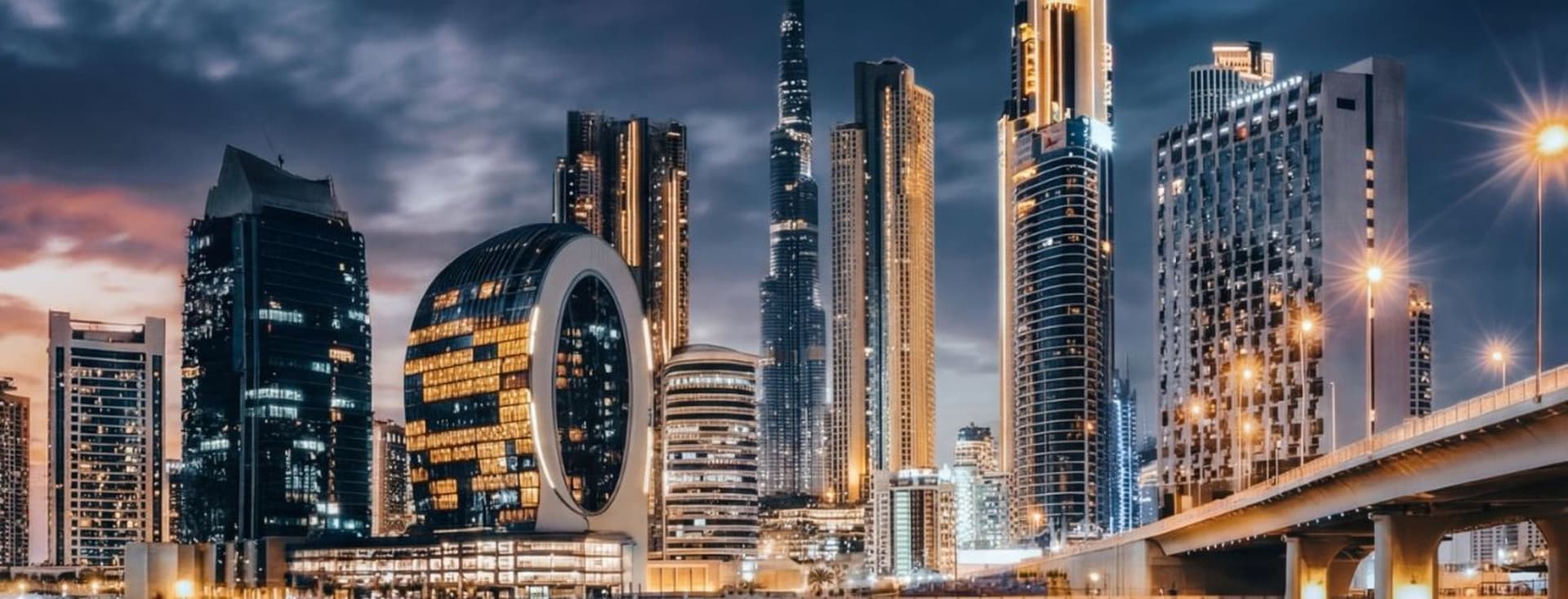 Dubai skyline at night with illuminated skyscrapers, including a distinctive circular building, reflected in calm water under a cloudy sky.