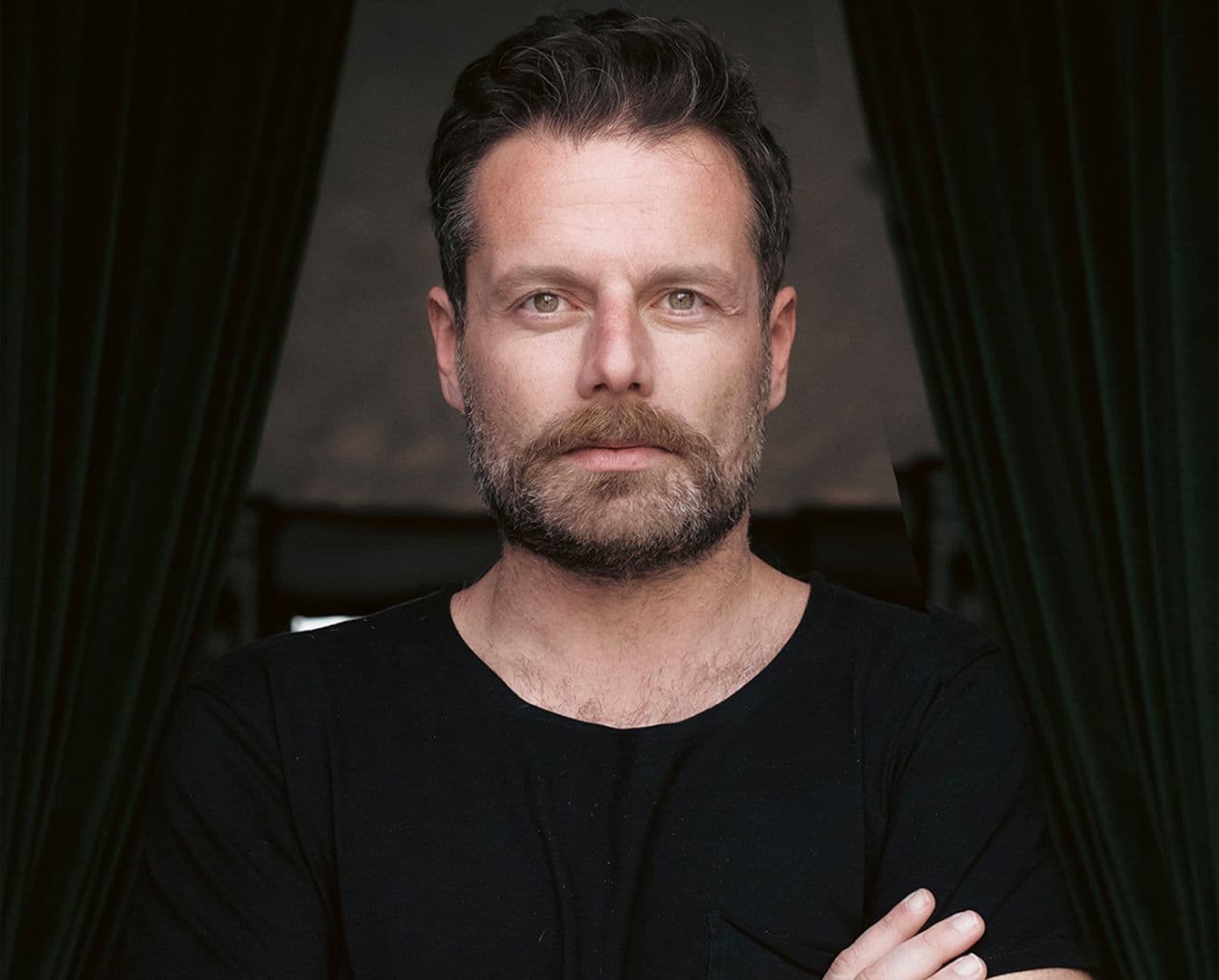 Porträt eines Mannes mit Bart, der ein schwarzes T-Shirt trägt, die Arme verschränkt, stehend vor dunkelgrünen Vorhängen.