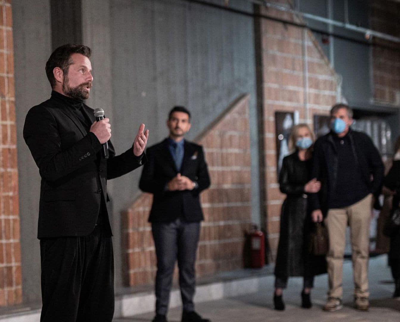 Ein Mann in einem schwarzen Anzug spricht in ein Mikrofon, während andere Leute im Hintergrund stehen.