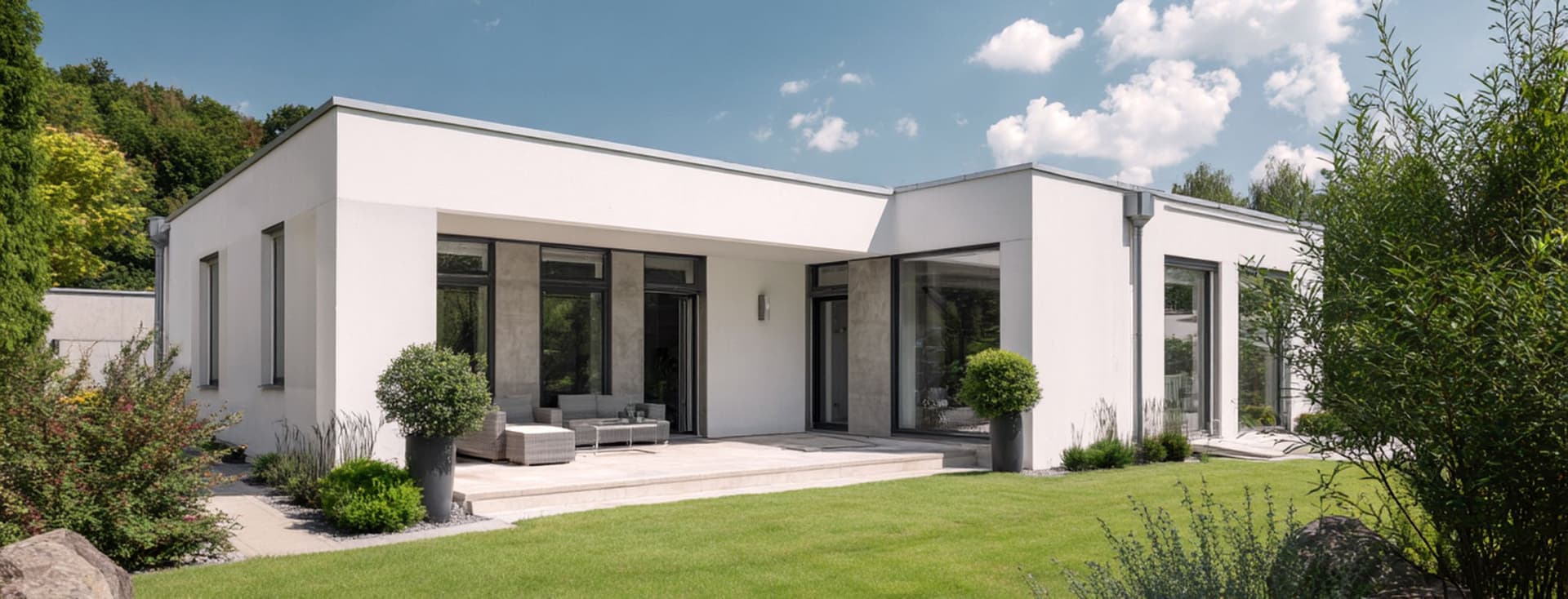 Modern white house with large windows, surrounded by a lush green lawn and shrubs, under a partly cloudy blue sky.