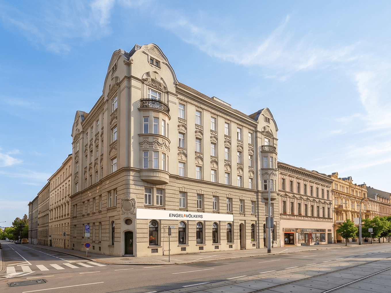View of a light-colored historic corner building with an ornate façade and bay windows housing the new Engel & Völkers branch in Brno. The building stands beside a wide road with tram tracks, surrounded by other city houses in a similar architectural style. Above the city is a clear blue sky with soft clouds.