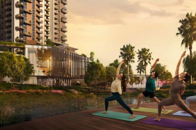Three people practice yoga on a wooden deck by a pond, with a modern high-rise building and palm trees in the background at sunset.