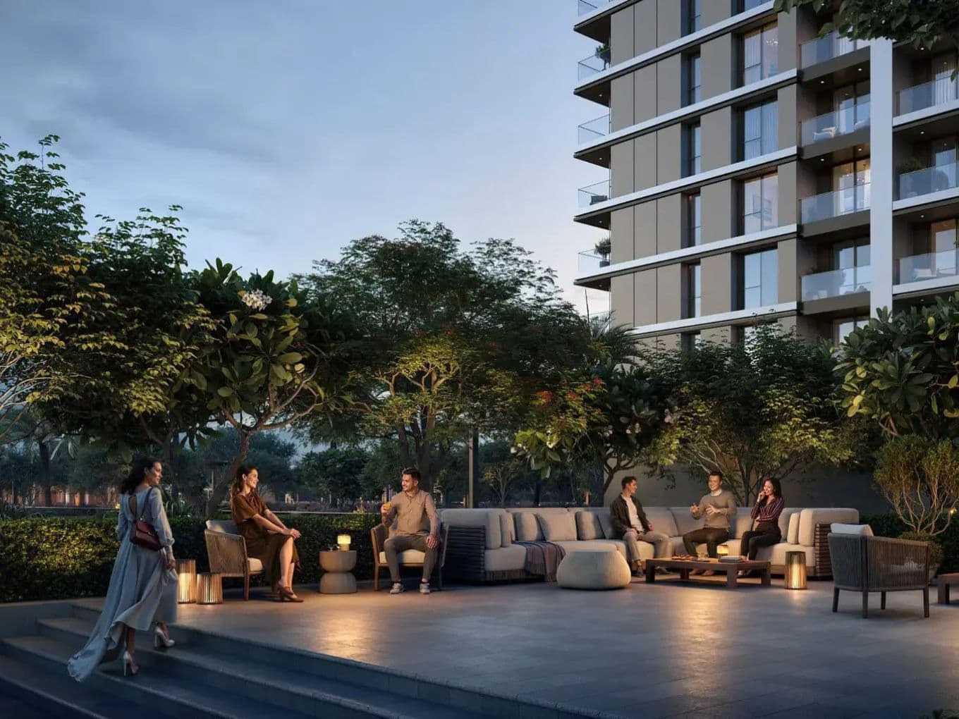 People sitting and talking on outdoor sofas near a modern building, surrounded by trees and illuminated by soft lighting at dusk.