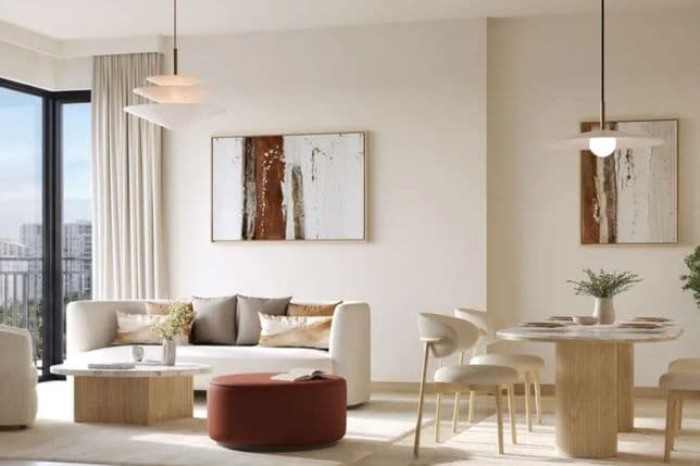 Modern living room with beige walls, abstract art, a white sofa, wooden tables, a rust-colored ottoman, and a dining area with pendant lighting.