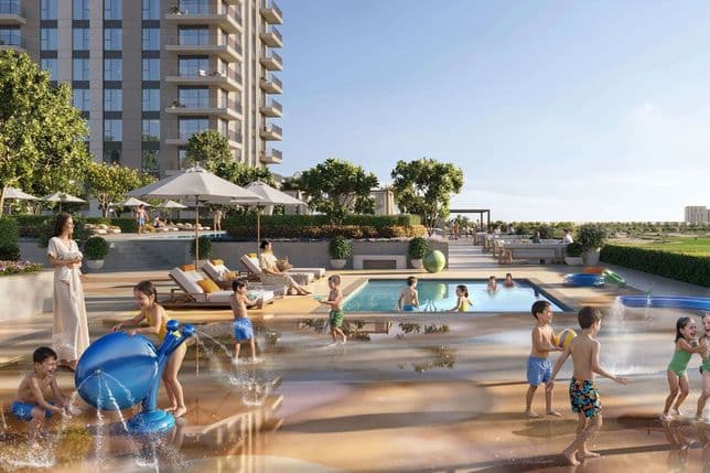 Children playing in a splash pad and pool at a modern apartment complex, with lounge chairs, umbrellas and high-rise building in background.