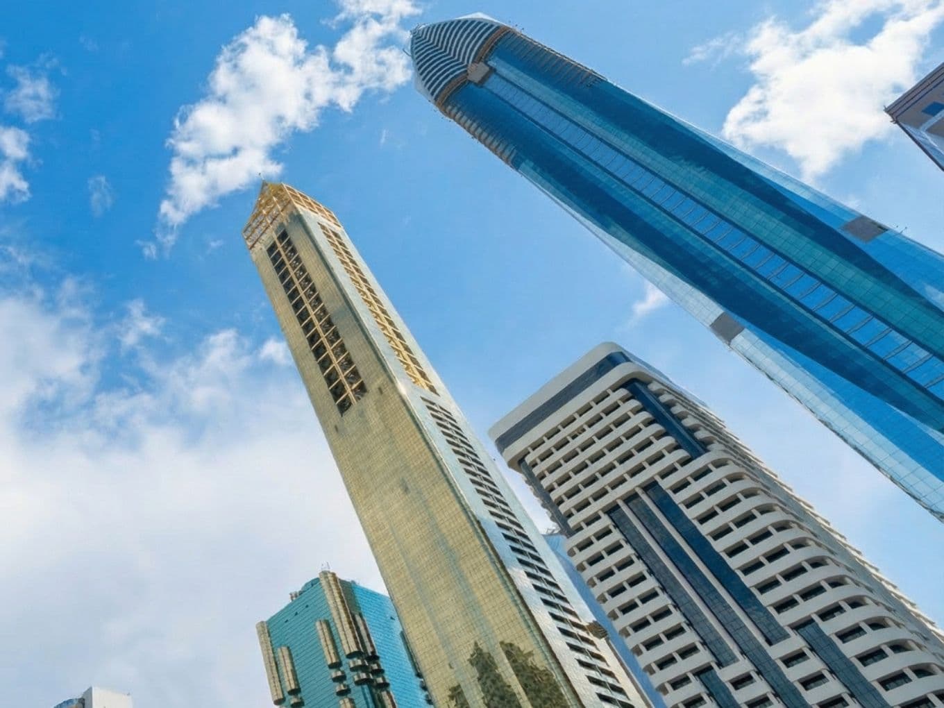 Tall modern skyscrapers with reflective glass facades against a bright blue sky with scattered clouds.