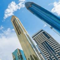Tall modern skyscrapers with reflective glass facades against a bright blue sky with scattered clouds.