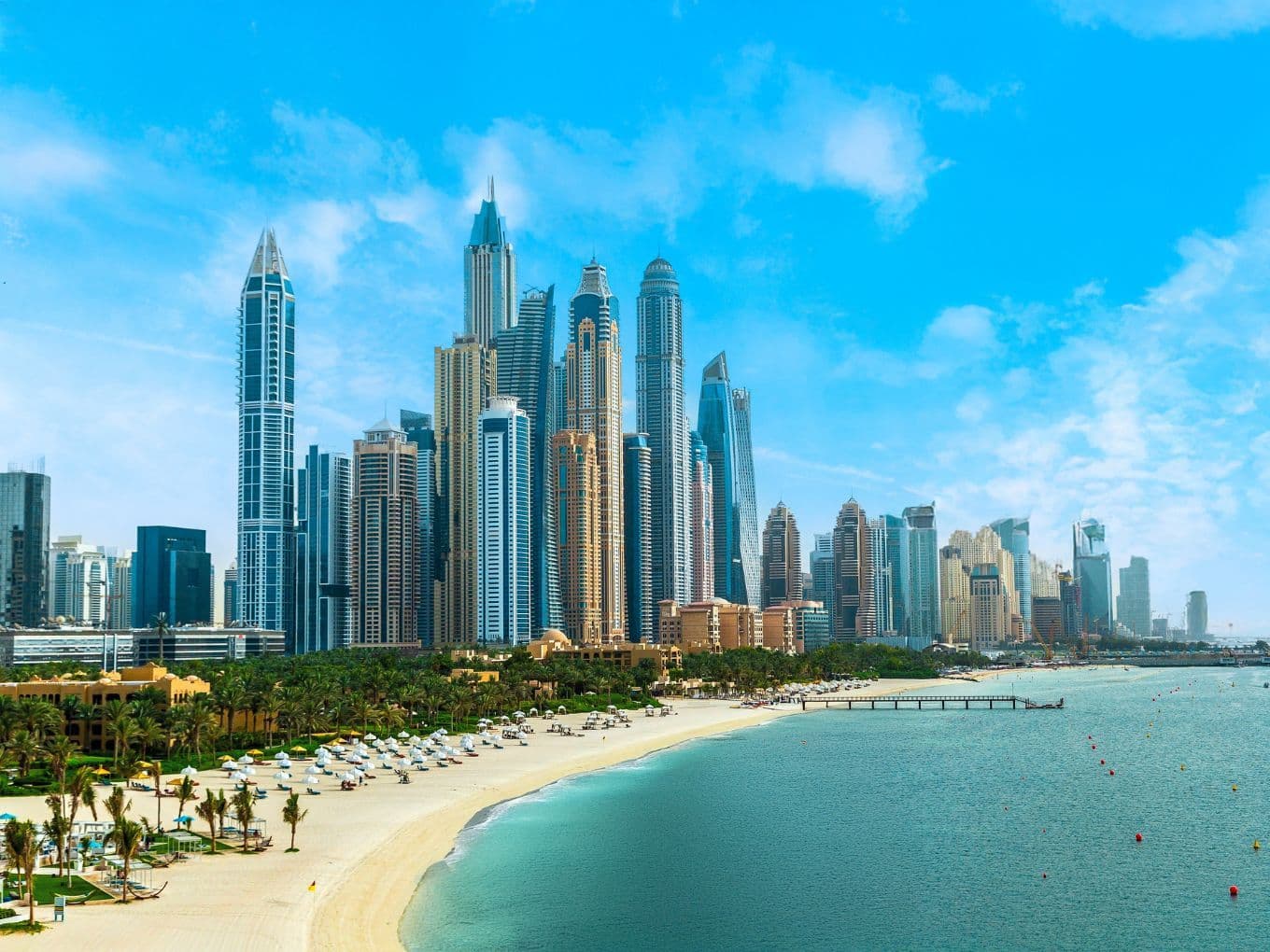 Skyline of Dubai Marina with numerous skyscrapers under a clear blue sky, adjacent to a sandy beach lined with palm trees and sun loungers.