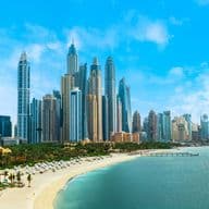 Skyline of Dubai Marina with numerous skyscrapers under a clear blue sky, adjacent to a sandy beach lined with palm trees and sun loungers.