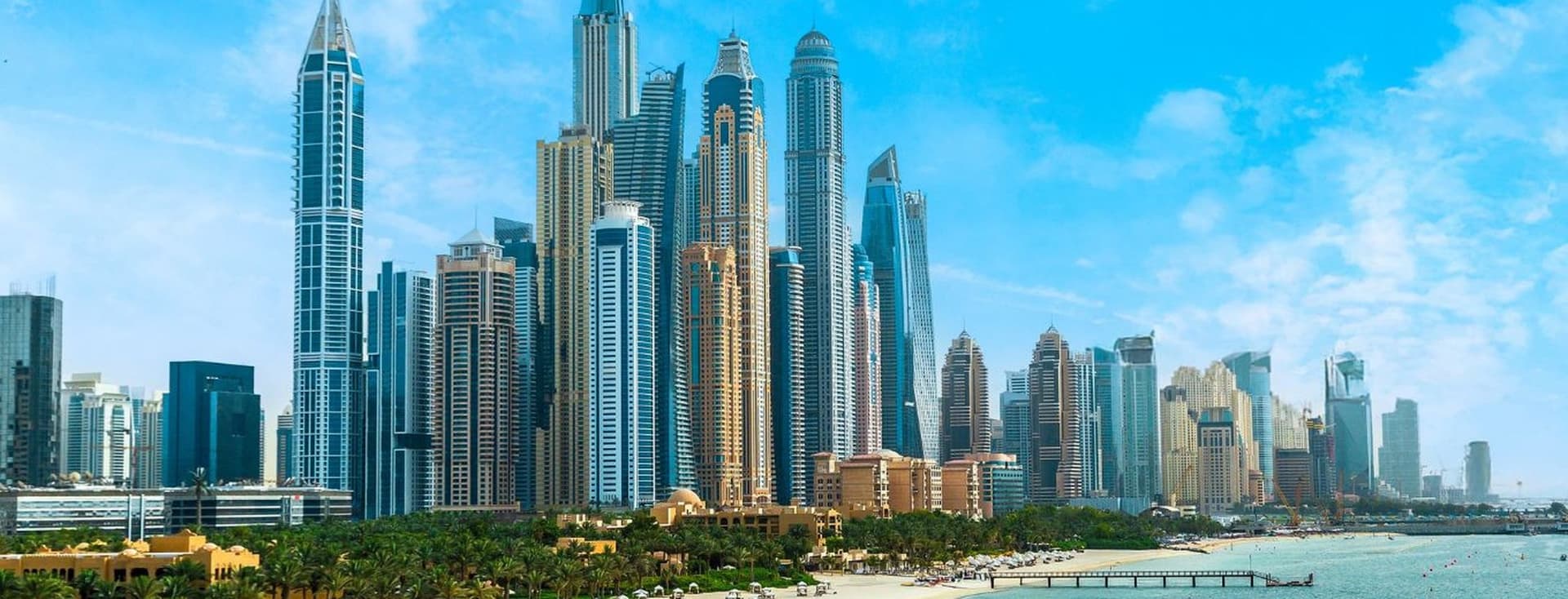 Skyline of Dubai Marina with numerous skyscrapers under a clear blue sky, adjacent to a sandy beach lined with palm trees and sun loungers.