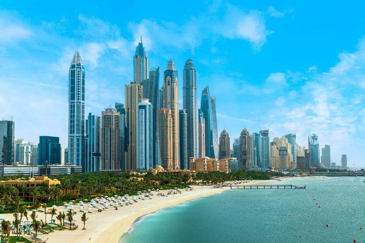 Skyline of Dubai Marina with numerous skyscrapers under a clear blue sky, adjacent to a sandy beach lined with palm trees and sun loungers.