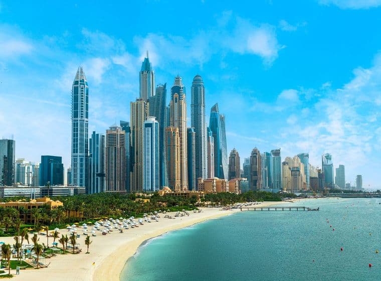 Skyline of Dubai Marina with numerous skyscrapers under a clear blue sky, adjacent to a sandy beach lined with palm trees and sun loungers.