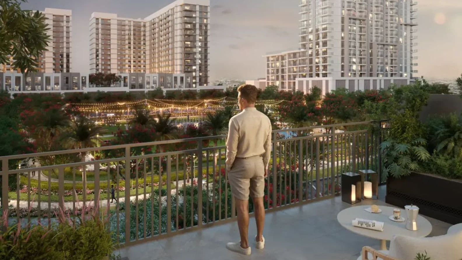 A person stands on a balcony overlooking a lush garden with string lights and tall modern buildings in the background at sunset.