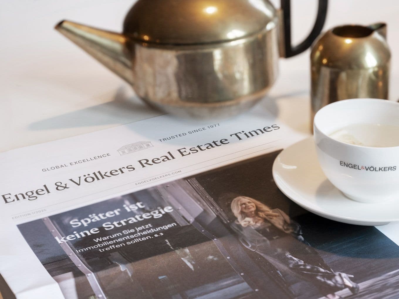 A newspaper titled "Engel & Völkers Real Estate Times" on a table with a teapot, small pitcher, and branded cup filled with liquid.