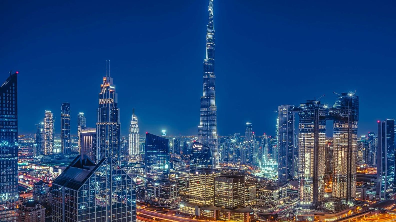 A vibrant cityscape at night featuring illuminated skyscrapers and winding highways, with a tall, iconic tower as the focal point.
