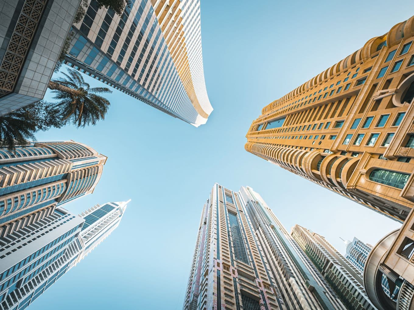 Upward view of modern skyscrapers against a clear blue sky, featuring diverse architectural styles and a palm tree in the corner.