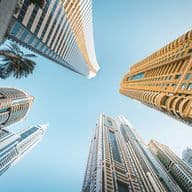 Upward view of modern skyscrapers against a clear blue sky, featuring diverse architectural styles and a palm tree in the corner.