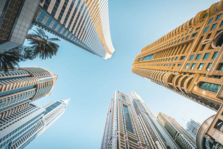 Upward view of modern skyscrapers against a clear blue sky, featuring diverse architectural styles and a palm tree in the corner.