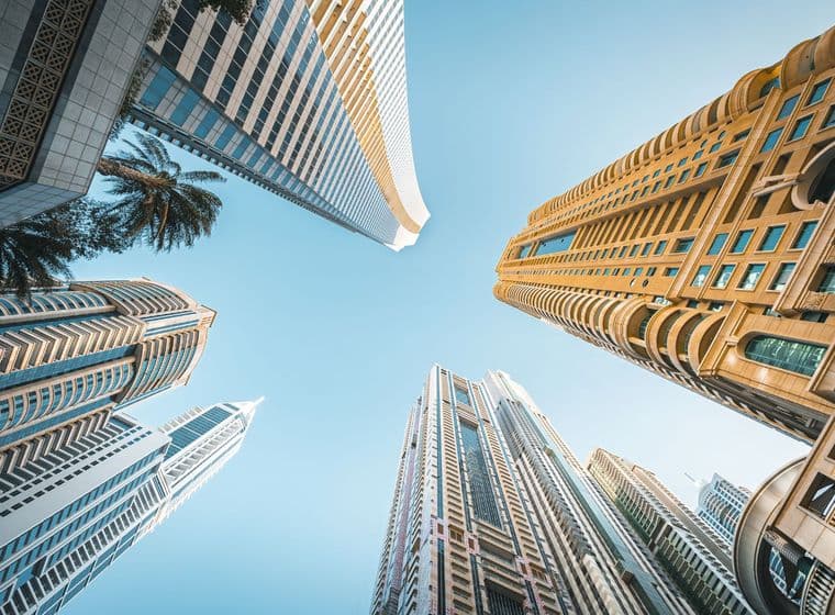 Upward view of modern skyscrapers against a clear blue sky, featuring diverse architectural styles and a palm tree in the corner.