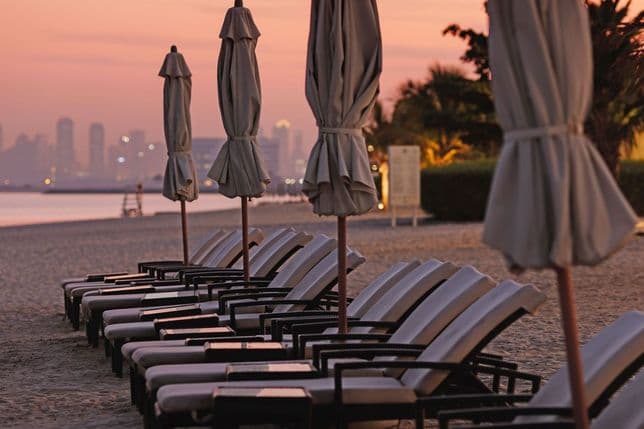 A row of beach lounge chairs and closed umbrellas on a sandy shore at sunset, with a city skyline in the background.