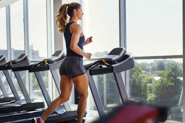 A woman runs on a treadmill in a bright gym with large windows, overlooking a cityscape with trees.