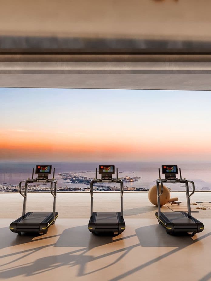 Modern gym with three treadmills facing a sunset over the sea through large glass walls; equipment lies on the floor, overlooking a calm horizon.