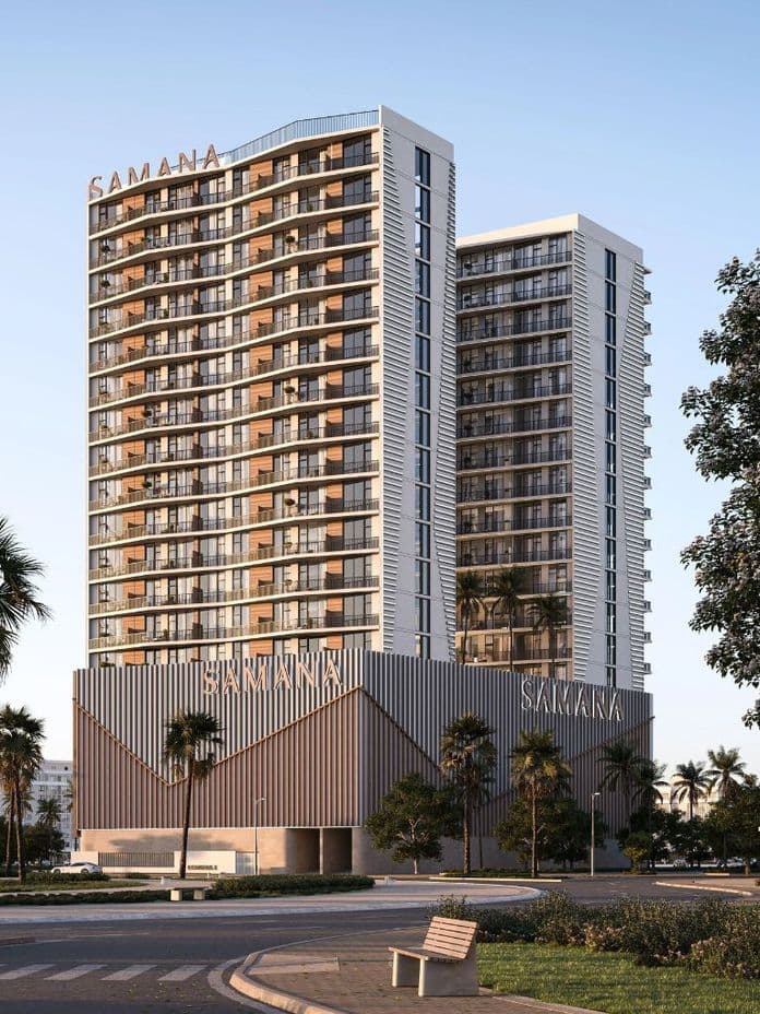 Two modern high-rise buildings with glass balconies, surrounded by palm trees, under a clear blue sky. A bench is in the foreground.