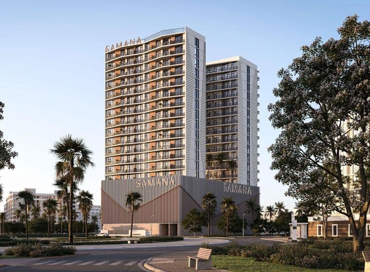 Two modern high-rise buildings with glass balconies, surrounded by palm trees, under a clear blue sky. A bench is in the foreground.