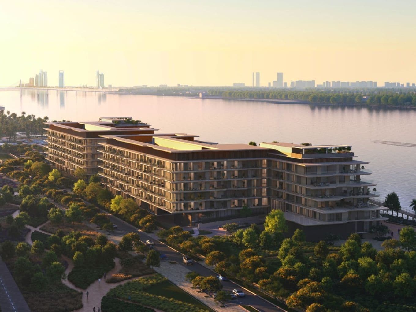 Sunset view of modern riverside apartment complex with balconies, tree-lined promenade and distant city skyline reflecting on calm water.