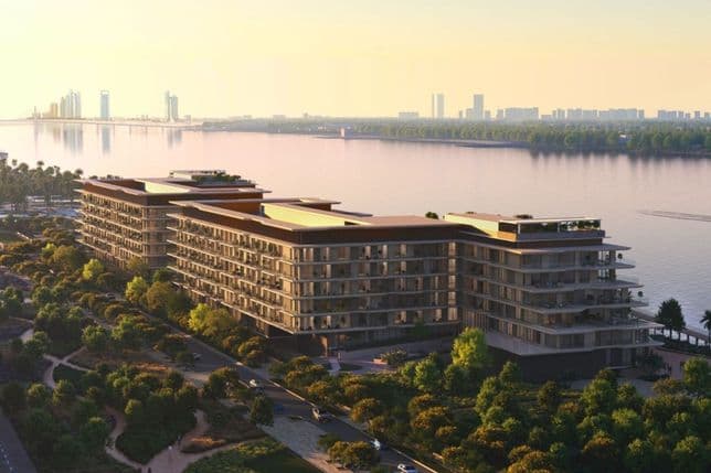 Sunset view of modern riverside apartment complex with balconies, tree-lined promenade and distant city skyline reflecting on calm water.