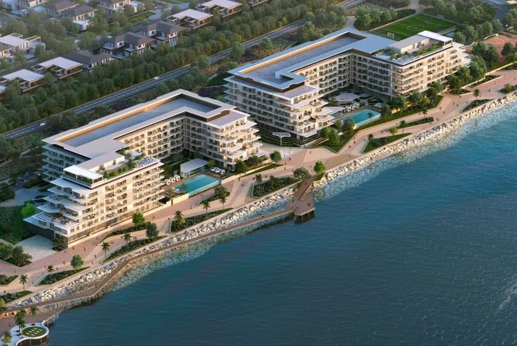 Aerial view of modern waterfront apartment complex with promenades, pools, greenery and a small pier along a rocky shoreline.