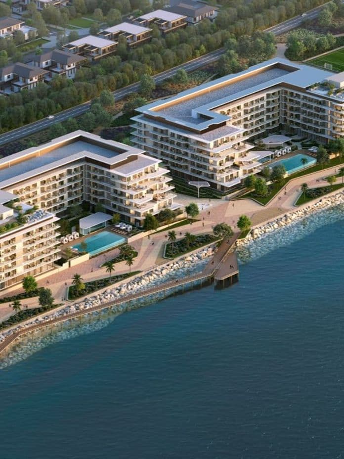 Aerial view of modern waterfront apartment complex with promenades, pools, greenery and a small pier along a rocky shoreline.