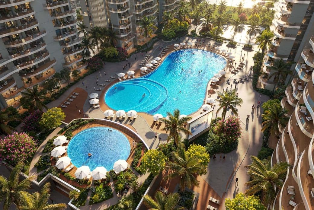 Aerial view of a modern apartment complex with two outdoor pools, surrounded by lounge chairs, umbrellas, palm trees, and gardens.