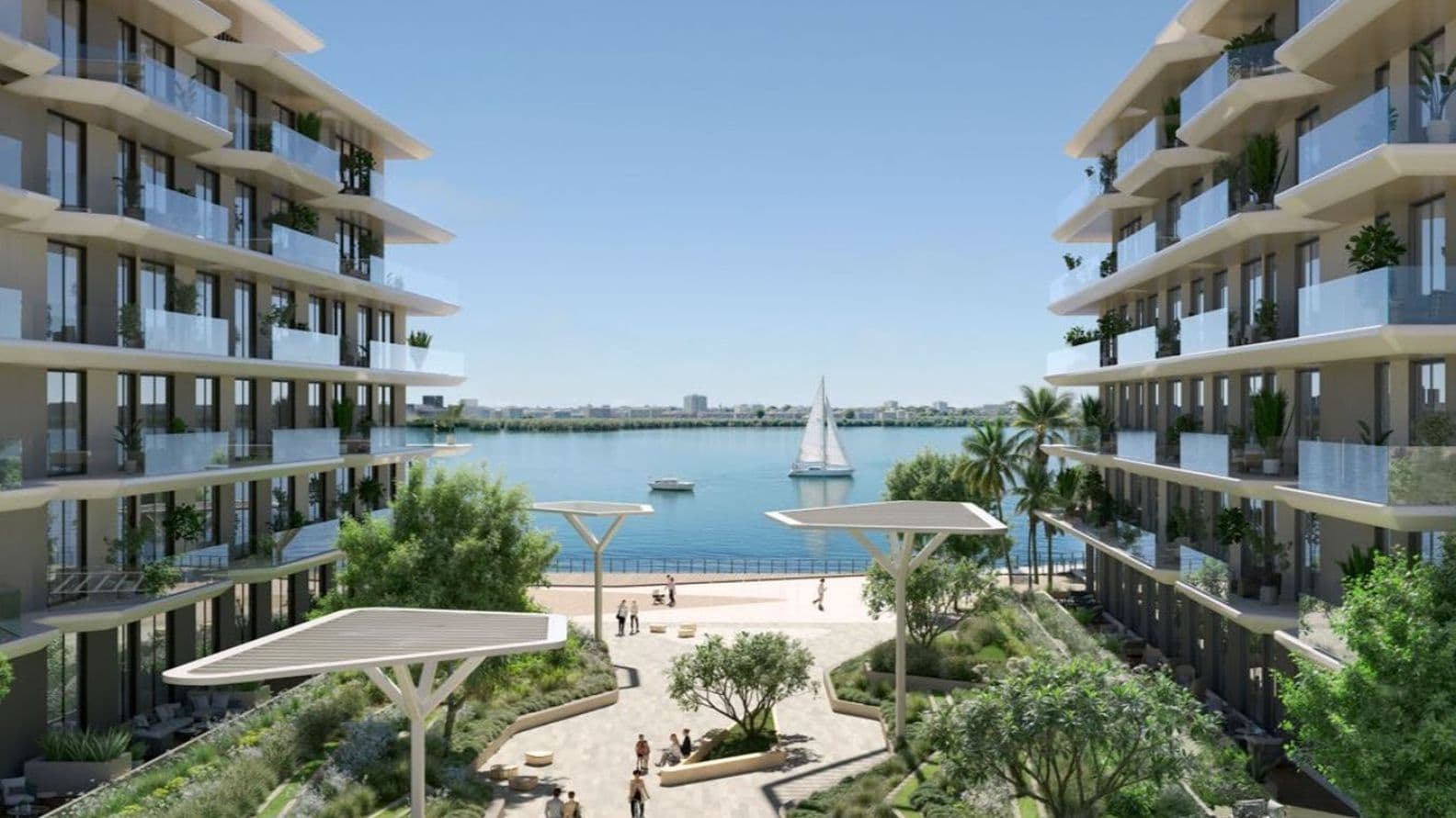 Modern waterfront apartment buildings flanking a landscaped promenade with people, palm trees and a sailboat on calm blue water.