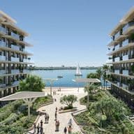 Modern waterfront apartment buildings flanking a landscaped promenade with people, palm trees and a sailboat on calm blue water.