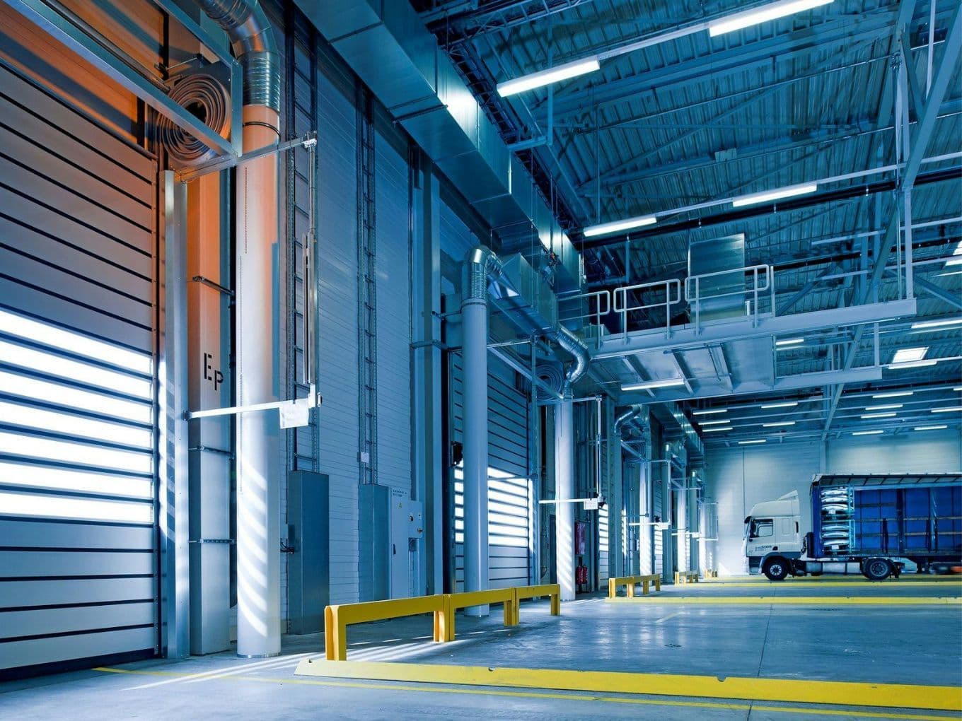 Spacious warehouse interior with high ceilings, industrial lighting, and a parked white truck near large bay doors.