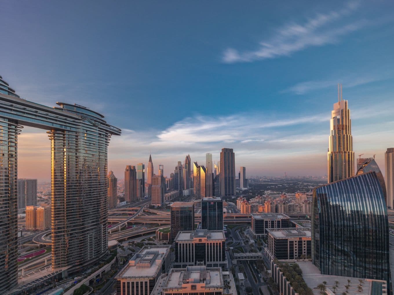 Skyline of a modern city at sunset, featuring tall skyscrapers, including a distinctive tower on the right, under a clear blue sky.