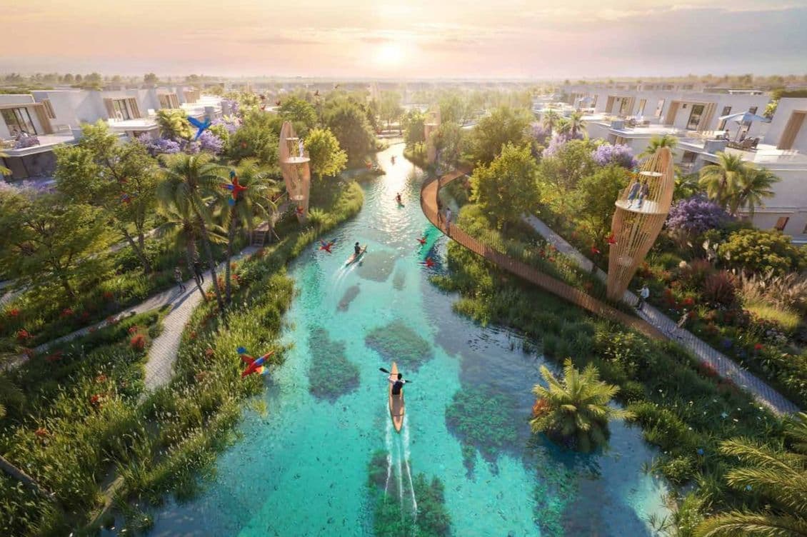 A serene canal winds through a lush modern residential area at sunset, with kayakers in the water and colorful birds in the trees.
