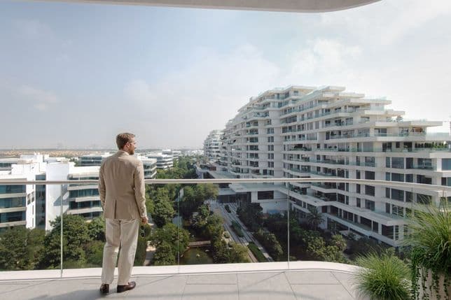 Eine Person in einem beigen Anzug steht auf einem Balkon und blickt auf moderne weiße Apartmentgebäude und viel Grün unter einem klaren Himmel.