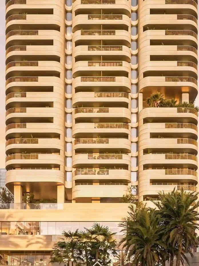 Modern high-rise building with curved balconies, lush greenery, and palm trees, set against a pastel sky in an urban environment.