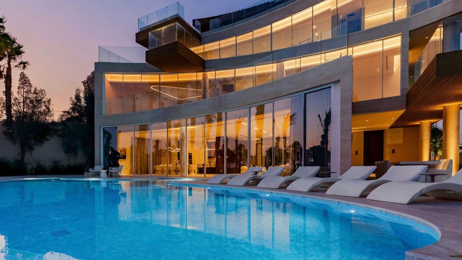 Modern luxury villa with large windows, illuminated at dusk, features a curved pool and sleek poolside loungers, surrounded by palm trees.
