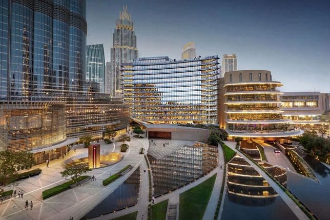 Modern cityscape with illuminated skyscrapers, reflecting pools, and a curved building at dusk. Paths and greenery surround the water features.