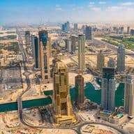 Aerial view of early Dubai Marina with skyscrapers under construction, winding roads, and a canal, set against a backdrop of sand and the distant coastline.